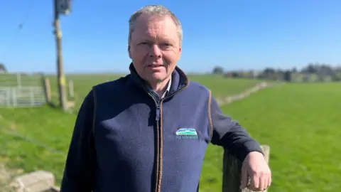 Andrew Connon, the president of NFU Scotland, leaning against a fence post on a farm. He has short grey hair and is wearing a blue fleece.