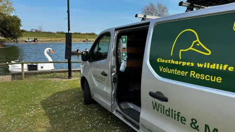 A silver van parked on grass facing a pond with a swan boat in. The van has yellow and green marking on, reading 'Cleethorpes Wildlife Volunteer Rescue'. The sky is blue. 