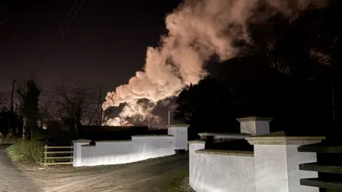 Plumes of white smoke rise from chimney stacks on Granville Industrial Estate at night against a pitch black sky. Front and centre of the image are two light grey plastered walls.