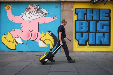 Glasgow to celebrate Sir Billy Connolly with weekend of events 3 Getty Images A mural of Billy Connolly wearing his "big banana boots". The caricature shows the comedian, naked from behind, with long white hair and a beard. "The Big Yin" is also painted on the wall. A man wearing black is walking past the mural, pulling a wheeled suitcase.