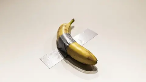 A piece of duct tape holds a banana to a white wall 