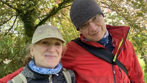 A man and a woman wearing hats are smiling at the camera. The man has his arm around the woman. She is wearing a baseball hat and the man is wearing a beanie. The woman has a blue and white scarf and is wearing a waterproof. The man has a red waterproof