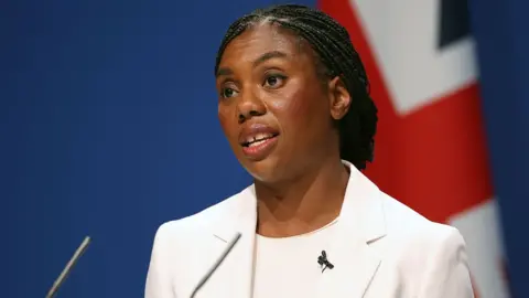 EPA Kemi Badenoch looks to the right of camera while wearing a white suit in front of a British flag against a blue backdrop as she talks into a microphone on stage.
