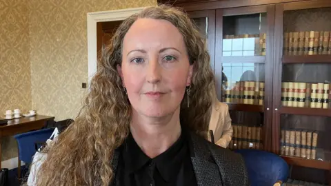 Rachel Hogan has light brown hair. She is wearing ear rings, a black shirt and a grey jacket. Behind her is a book shelf with wooden and glass doors, there are books inside on shelfs. There are blue chairs and a side table in the background. There is also a wooden door, with a white door frame behind her. 