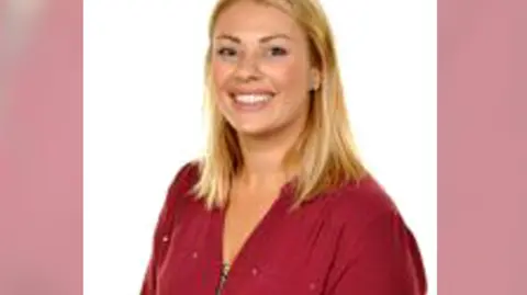 A woman smiling at the camera. It is a head and shoulders shot. She has blonde hair and a red top on.