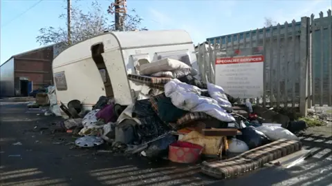 Fly-tipping rubbish, including mattresses and bedding, piled up against an old white caravan