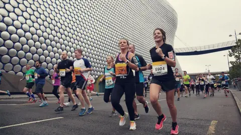 Male and female runners running past Selfridges building in Birmingham city centre. 
