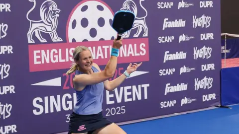 Thaddea is in a blue vest and blue skort, with her racket in the air, backed by a purple sponsorship board.