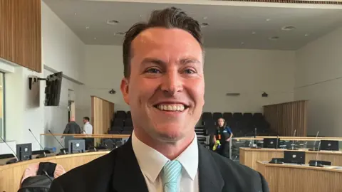 Smiling white man with brown hair. You can see the top of a suit and tie and figures in the background along with wooden podiums and empty seats