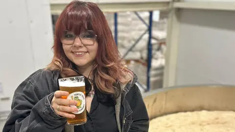 Imogen-Rose Gregory raises a pint of beer alongside a vat of hops