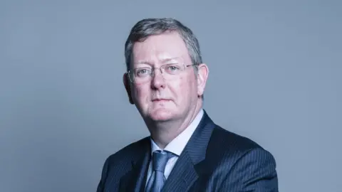 UK Gov Lord Caine with short dark hair wearing a black striped suit, grey tie and white shirt as he stares at the camera.