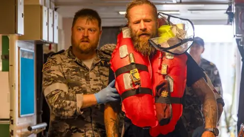 Australia Defence Force Chief Petty Officer Medic Peter Gough in military gear leads solo rowboat adventurer Aurimas Mockus, who is in an orange life jacket, to a hospital onboard the Australian warship HMAS Choules