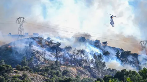A helicopter drops water on a wildfire that rapidly expands due to strong winds near the city of Marseille, France, 08 July 2025.