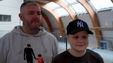 Father and son Steve and Ollie stand next to each other. Steve has short, cropped grey hair and a beard. He is wearing a grey hoodie. His son Ollie wears a black baseball cap and a black round-necked top.
