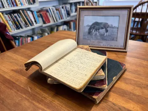 An open book with handwriting sits on top of three closed books on a table. In the background is a framed photograph of a horse and foal, with the inscription Vic, 1920.
