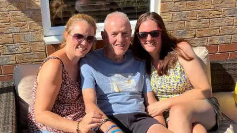 Three people sit closely together on a cushioned outdoor wicker sofa in a sunlit patio area. A brick house wall with a window is directly behind them. A small table in the foreground holds a takeaway coffee cup and other items. The group is dressed casually in warm-weather clothing.