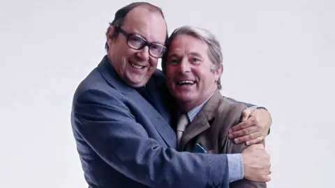 Close up of Eric Morecambe with his arms around Ernie Wise giving him a big hug. Both are smiling, Eric is wearing a blue suit, has receding dark hair and dark-framed glasses. Ernie is in a brown suit and has short white hair