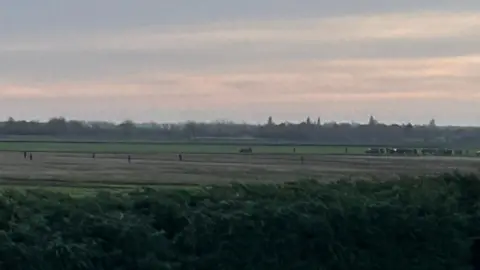 Mathew Latta From a distance, a gang of hare coursers can be seen walking and driving through a field. The sun is setting and it is getting dark. 