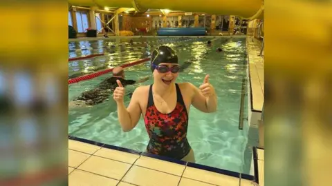 Journalist Eleanor Maslin in a speedo costume with stars on putting both her thumbs up at the end of a pool whilst wearing her goggles and Pudsey swim hat. Lanes and other swimmers can be seen behind her.