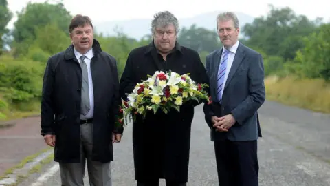 Pacemaker Three men standing side by side as one holds a wreath. From left to right is a man with short brown hair black coat, grey trousers, white shirt and grey tie. then a man with short grey hair and a black coat. Then a man with short grey hair, grey moustache wearing grey jacket, white shirt, blue striped tie and black trousers. Behind them is a road where the attack occured.