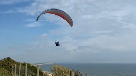 AndrewP A paraglider floats over the coastline with the sea behind him and a beach trailing into the distance
