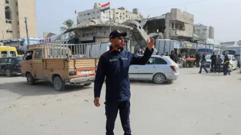 Reuters A Hamas police officer directs traffic in Gaza City. He's blowing an orange whistle while wearing a blue cap and a blue uniform. A white car and a brown truck drive behind him. 