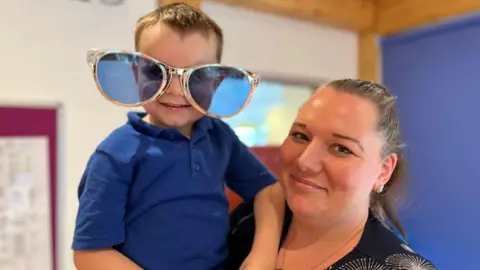 A woman holds a young boy. She is wearing a black and white blouse. He is wearing a blue t-shirt and novelty oversized plastic sunglasses with gold frames.