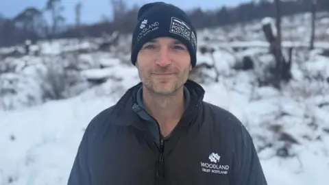 Liam Wright is a smiling, dark-haired man in a blue Woodland Trust Scotland hat and jacket.