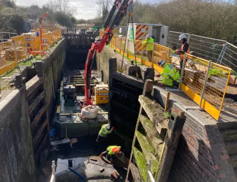 Canal and River Trust Image shows the right new-look gate being lifted into the air by a small crane and held up by two workmen as it is put in place.