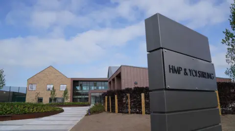 A general view of HMP and YOI Stirling shows a modern, blonde-bricked two-story building with new landscaping surrounding it and a sign with " HMP and YOI Stirling " in large black letters.