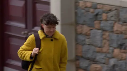 Deputy Tina Bury - A lady wearing a yellow wooly coat, with a bag strap over her right shoulder, walking up the road. She has curly brown hair on the top of her head and short hair on the side. 