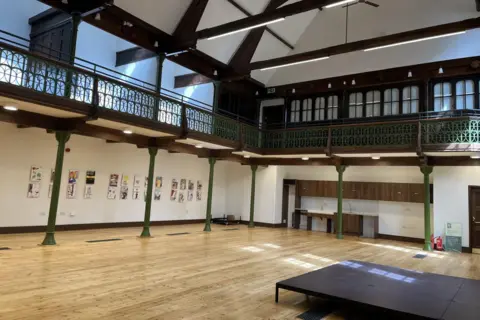 Inside the Sunday School hall with stage on the wooden ground floor and a balcony standing above green metal pillars