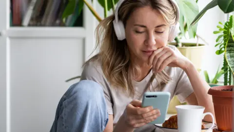 A stock shot of a young blonde woman wearing white headphone and looking at her phone.