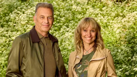 BBC Studios Chris, left, is looking at the camera. He is wearing a khaki green bomber style jacker and a green polo top. He has short grey hair. Michaela, right, is wearing a beige zip up coat and a green t-shirt underneath. She has shoulder length blonde hair with a fringe. She is smiling at the camera.