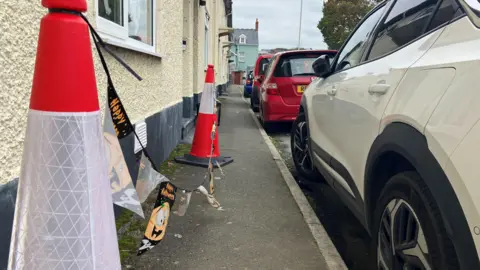 Two orange traffic cones on a street pavement, with bunting reading 'happy halloween' attached to them. 