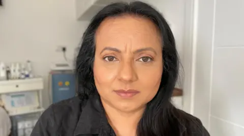 BBC Sukhjeet Bahra looks directly at the camera. She has dark black hair and is wearing a black shirt with a collar. 