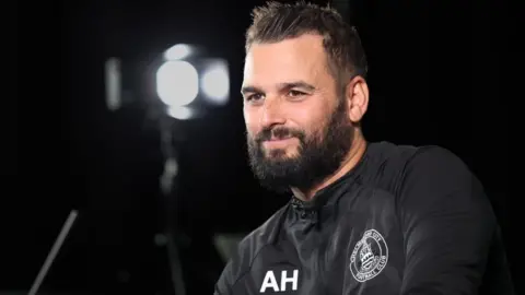 Angelo Harrop, wearing a Chelmsford City black tracksuit top, sitting for down with an interview with lighting in the background.