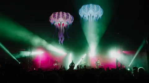 Andy Holbrook Fans applaud Radio Free Alice as they take to the Giant Squid stage. There are fluorescent lights going into the crowd. 