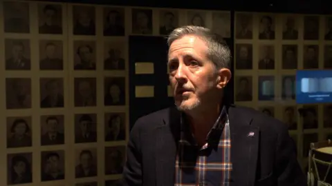 Vic Tanner-Davy is wearing a black blazer and is standing in front of a wall of pictures