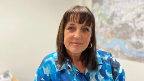 A woman with shoulder length dark hair and a fringe smiling at the camera. She is wearing a blue blouse and there is an out of focus map behind her.