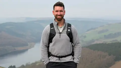 Sam Culley Sam Culley wearing black shorts and a grey pullover, with a black rucksack strapped across his chest and waist. He has short brown hair and a beard, and is smiling at the camera with his hands in his front pockets. Behind him there is a sweeping view of a lake, hills, fields and woodland on a misty day.