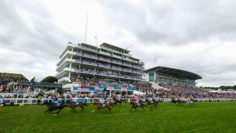 Blurred horses pass the winning post in a race at the Epsom Derby festival with stands full of spectators visible in the background