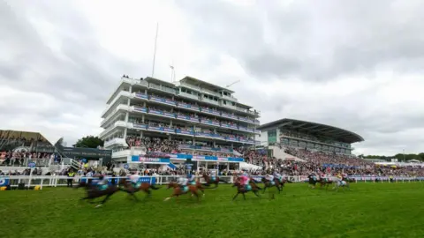 Getty Images Blurred horses pass the winning post in a race at the Epsom Derby festival with stands full of spectators visible in the background