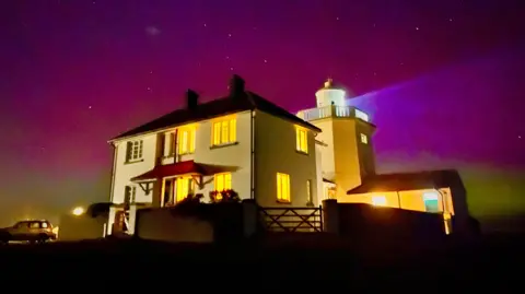 BBC Weather Watchers/Walking Tractor A white house and lighthouse is in the foreground. The Northern Lights have made the sky turn purple, pink, and yellow, and there are several stars.