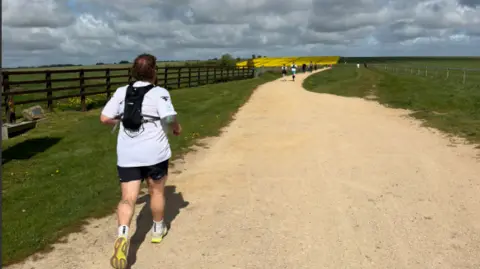 Way Swindon A man running down a dirt path with green and yellow fields in front of him. The sky is very cloudy. There are people in the distance running ahead of him.