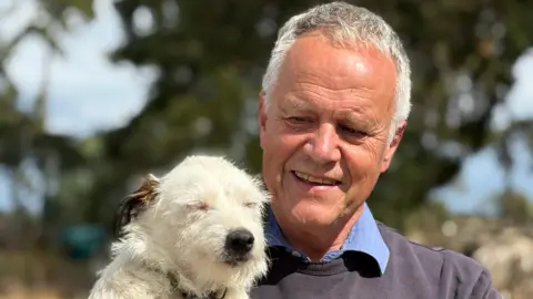 A man with grey hair wearing a dark grey jumper holding a white Parson Russell Terrier