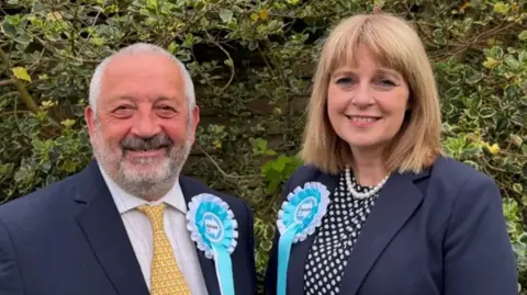 Marcus Lapsa and Jackie Gardiner look at the camera. They are wearing dark blue blazers and light blue and white Reform UK rosettes.