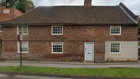 Google An 18th Century steep roofed building with small windows and a light blue door sits in front of a footpath and strip of grass