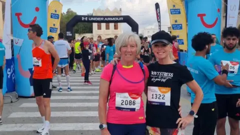 Nicky Putman Two women stood at the finish line of a race. One is wearing a pink top, the other in black with both having race numbers on their chests. They have their arms around each other and are smiling at the camera. There is TUI branding around them along with lots of other runners. 