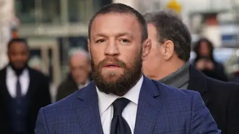 PA Media Conor McGregor mengenakan kemeja biru, kemeja putih, dan dasi hitam. Dia memiliki rambut pendek berwarna coklat muda dan janggut